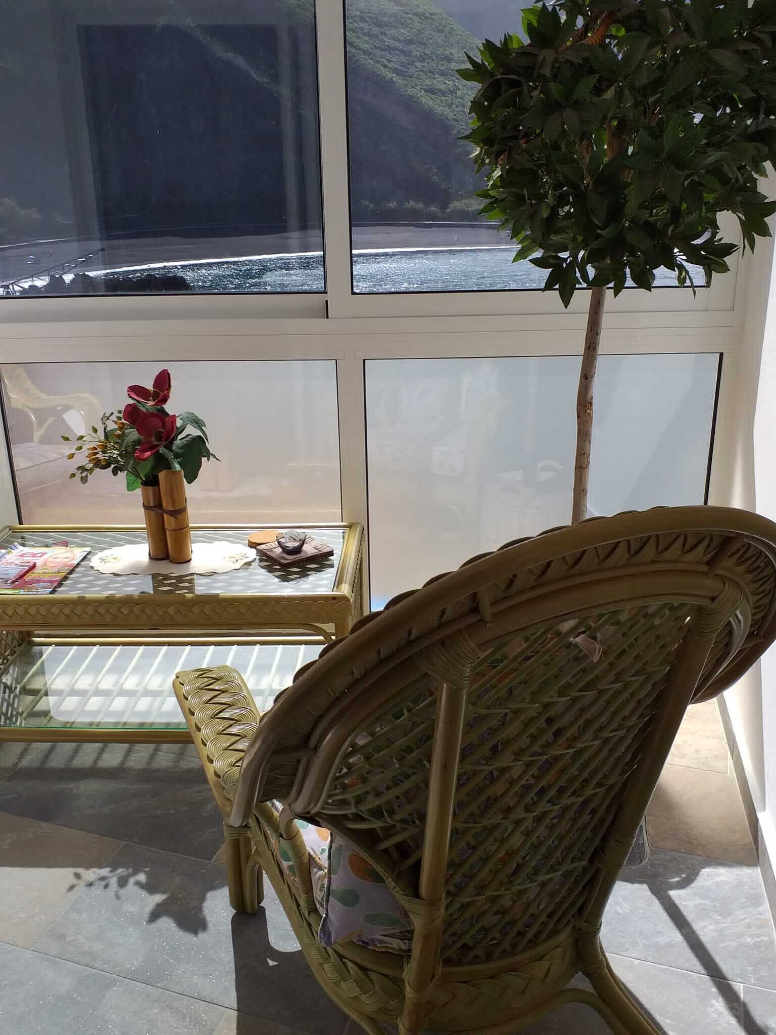 Una habitación con un ventanal amplio, una silla de mimbre, una mesita con un florero y una planta en maceta.