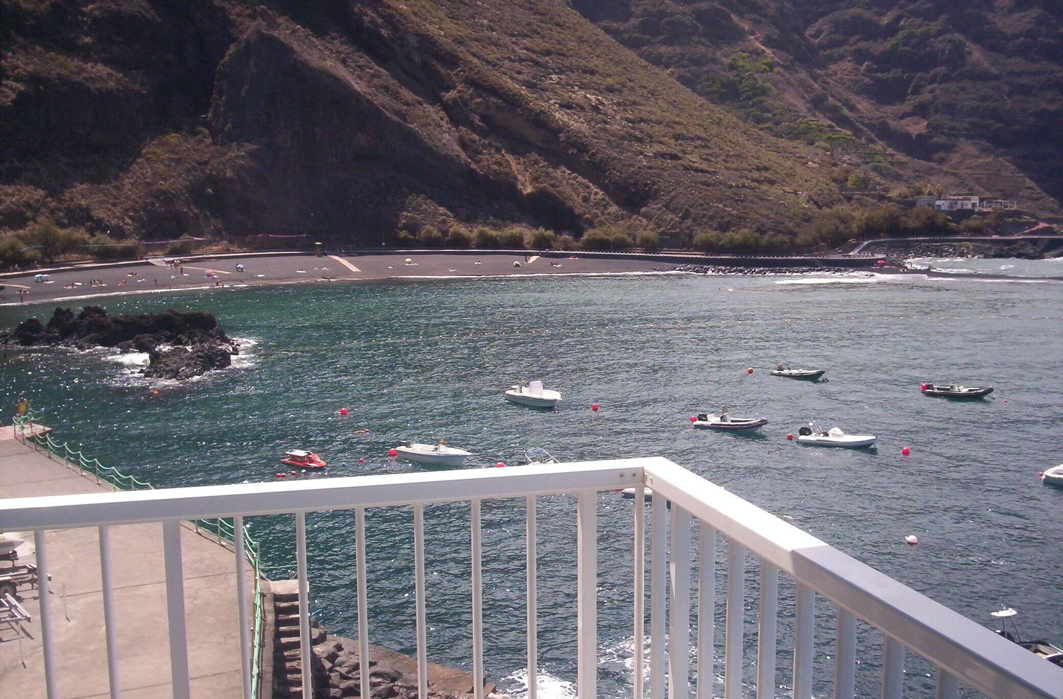 Zona costera con barcos en el agua, una carretera con vehículos y montañas al fondo.