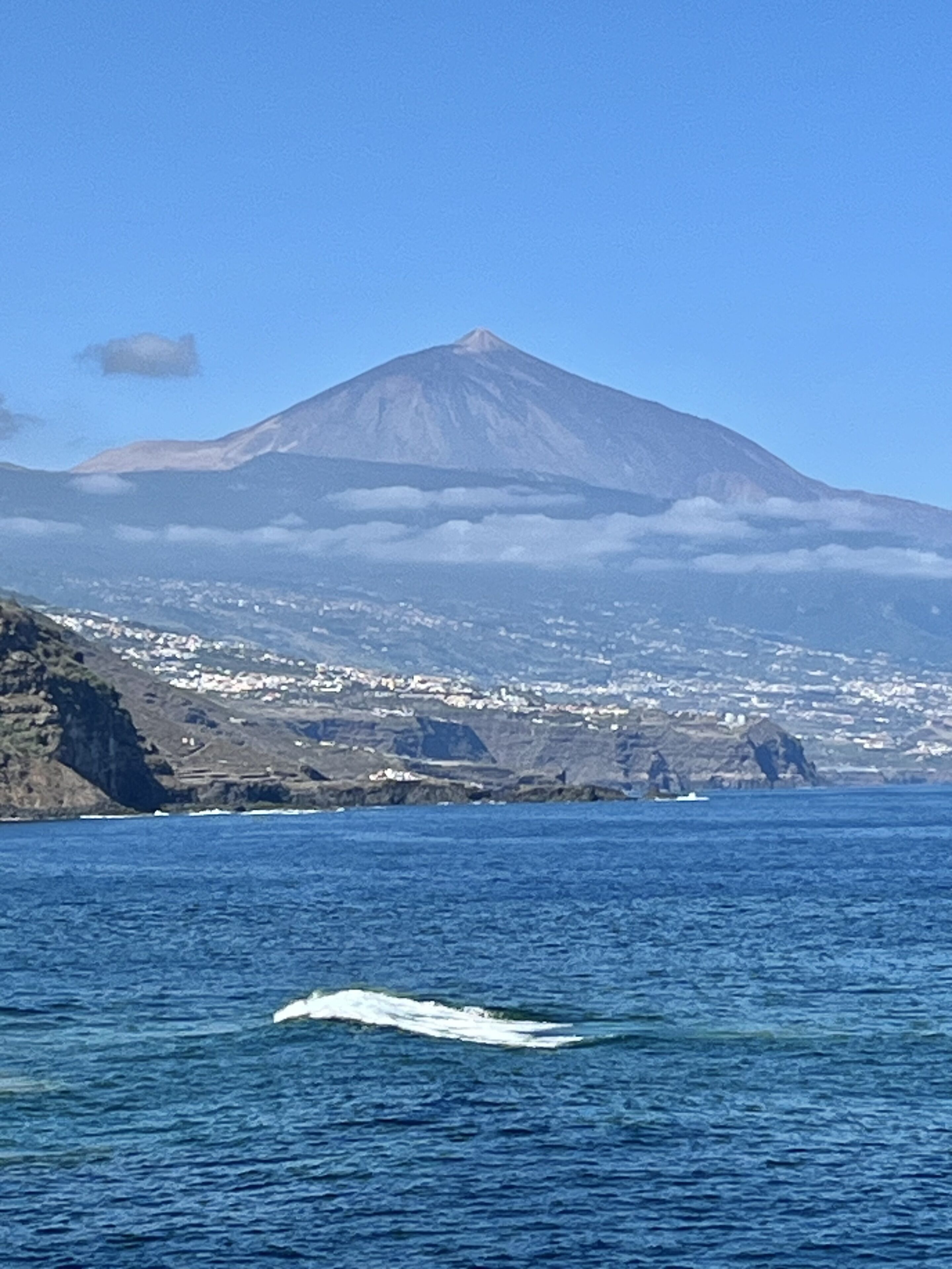 Un paysage côtier avec une montagne imposante, un petit bateau sur l’eau et une côte lointaine parsemée de bâtiments.