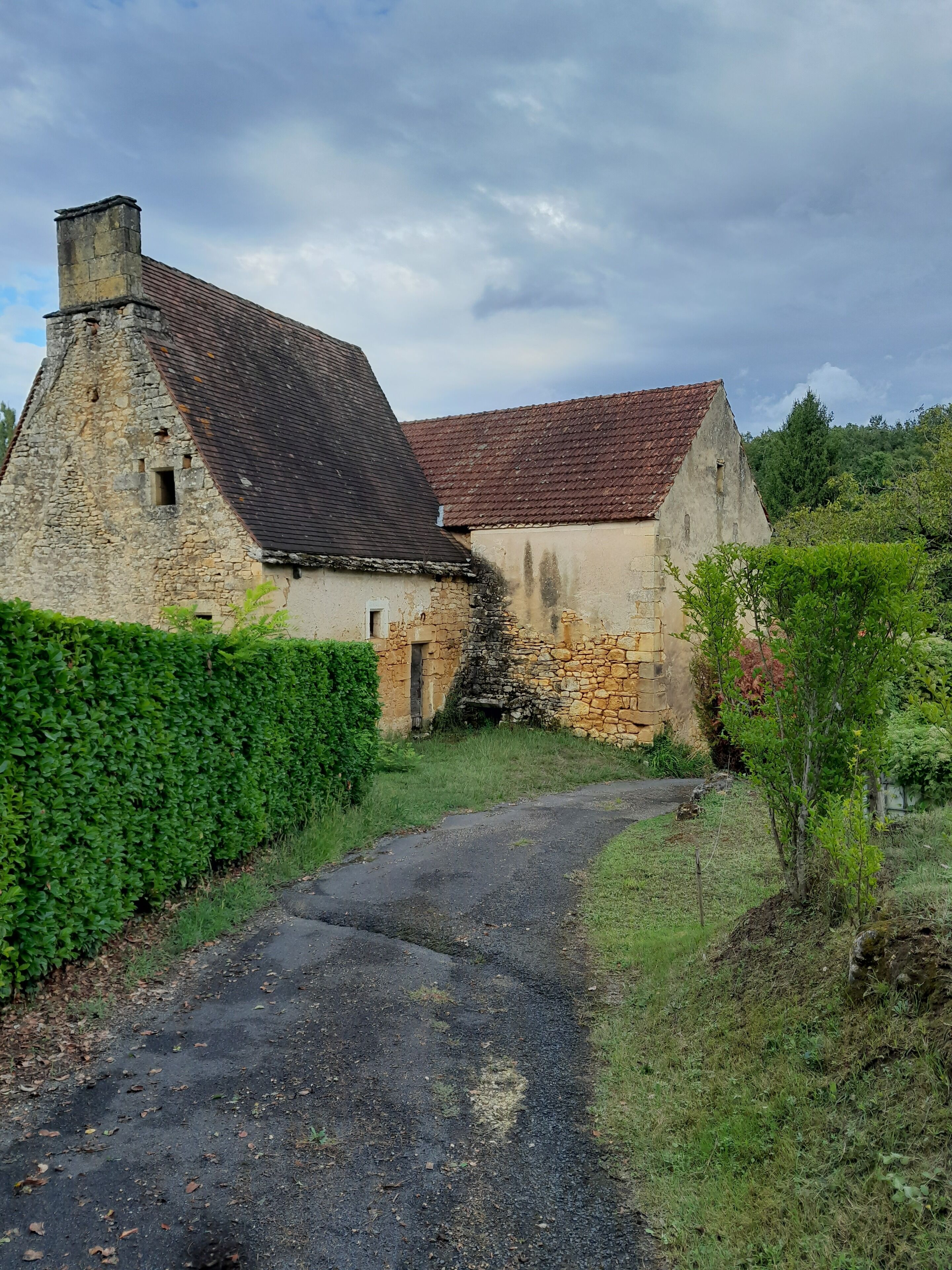 A rustic stone building with a sloped roof, a narrow pathway leading to the entrance, and a well-maintained hedge on one side.