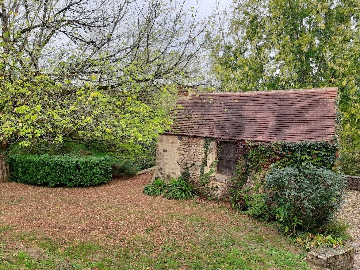 A rustic stone cottage with a sloped roof, surrounded by dense greenery and trees.