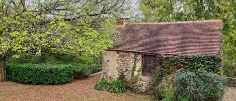 A rustic stone cottage with a sloped roof, surrounded by dense greenery and trees.