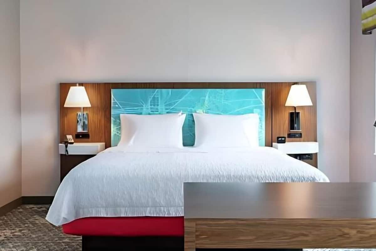 Une chambre d’hôtel avec un grand lit, deux tables de chevet avec des lampes, une tête de lit en bois et un coussin décoratif rouge.