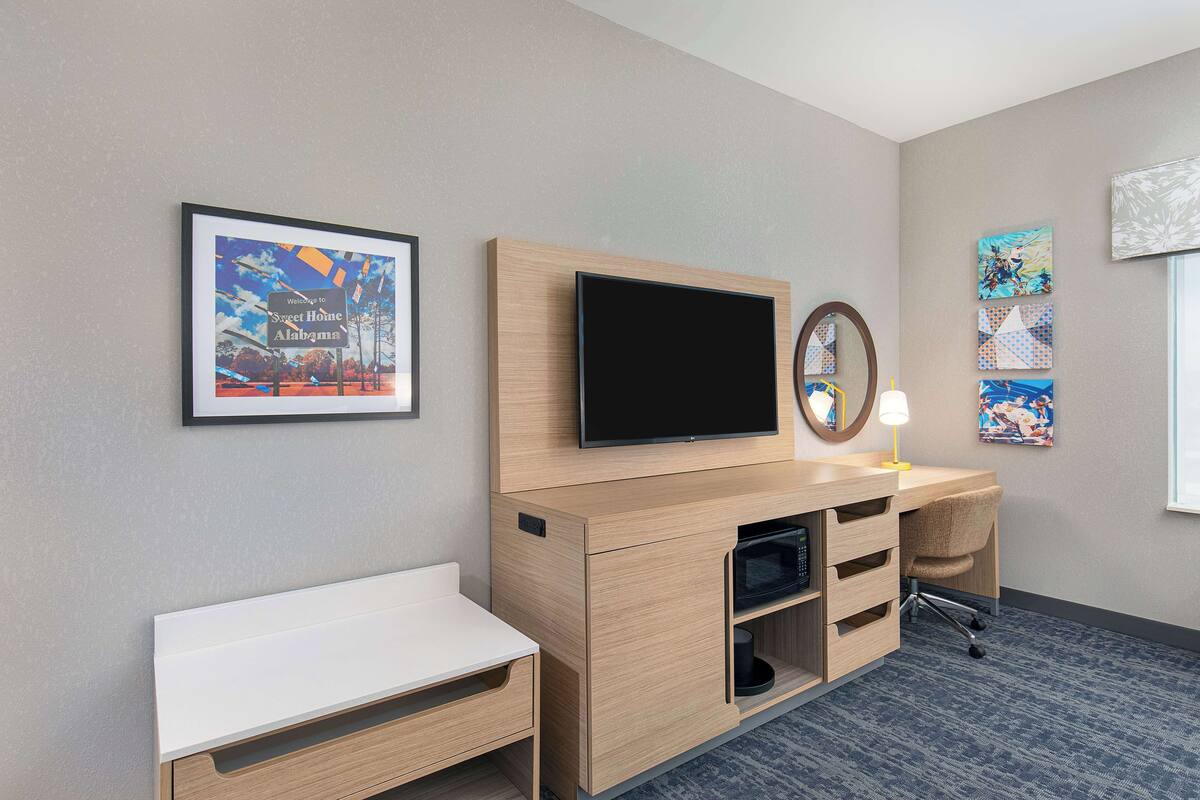 Une chambre d’hôtel dotée d’un meuble de rangement en bois, d’un téléviseur à écran plat, d’un bureau avec une chaise et d’œuvres d’art accrochées aux murs.