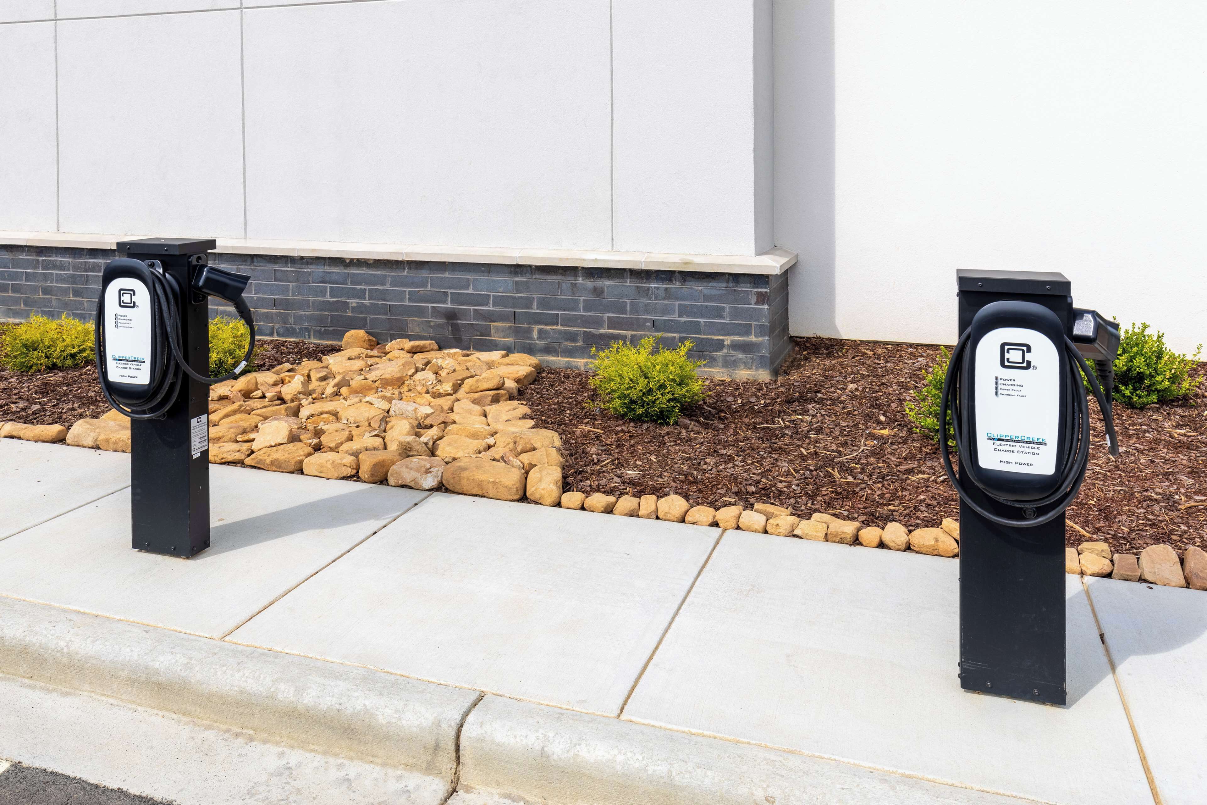 Two electric vehicle charging stations with labels, set against a backdrop of a building and a landscaped area with rocks and small shrubs.