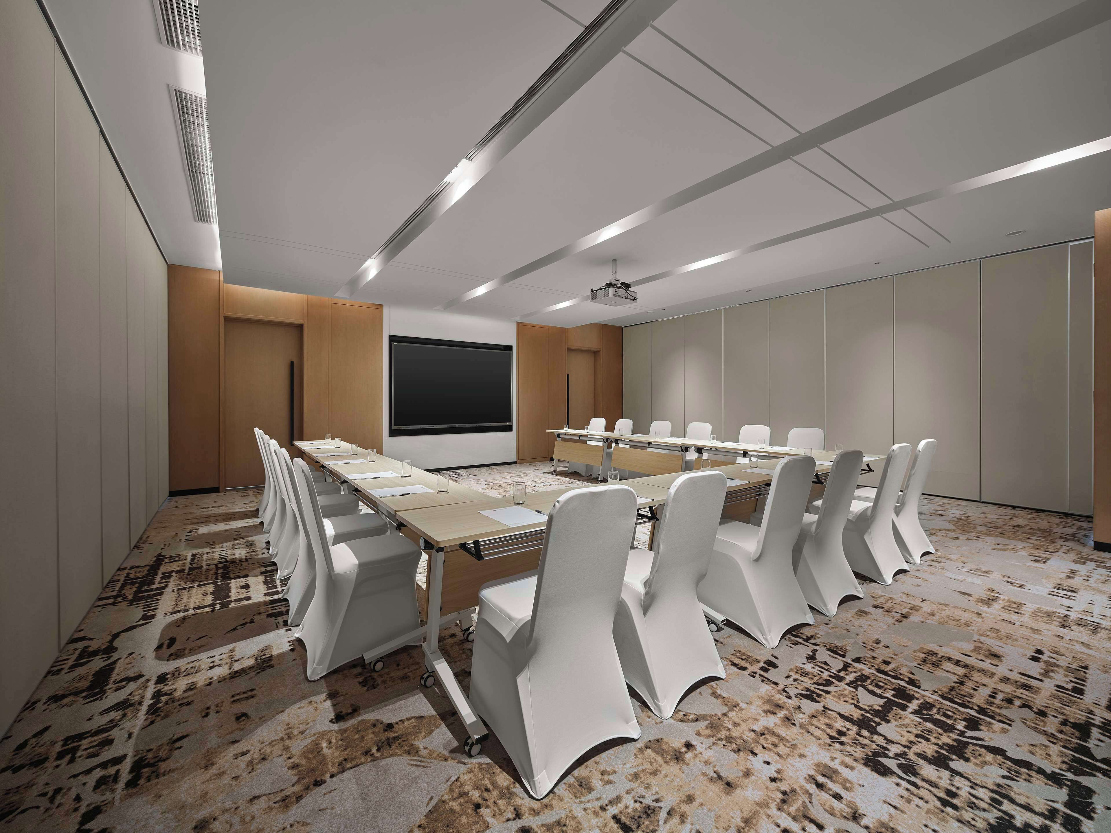 A conference room with a long table, white chairs, a projector screen, and a carpeted floor.