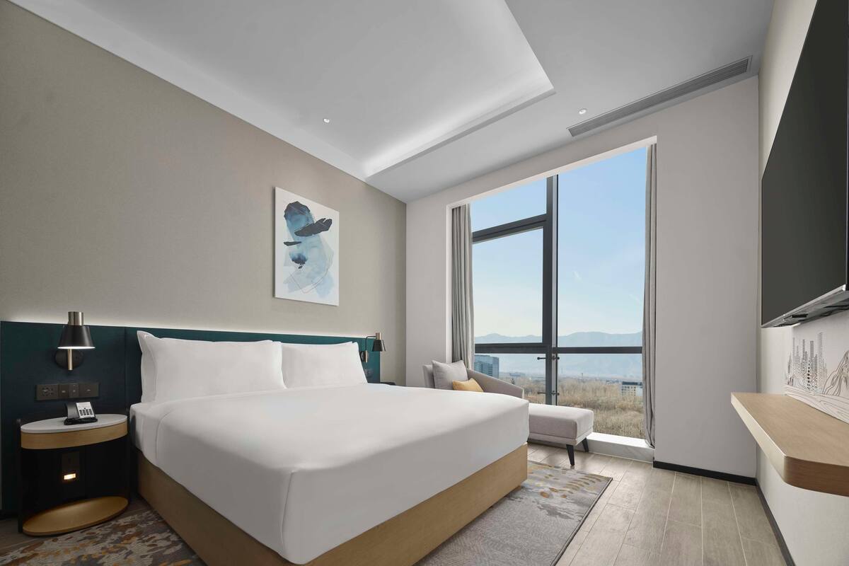 Habitación de hotel moderna con una cama grande, un escritorio, una silla, un cuadro y vistas a las montañas.