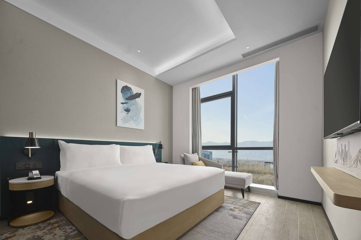 Una habitación de hotel moderna con una cama grande, una mesita de noche con lámpara, vistas a las montañas a través de la ventana y una vista de la ciudad en la pared opuesta.