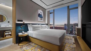 Una habitación de hotel moderna con una cama grande, un baño con espejo y vistas a la ciudad a través de amplios ventanales.