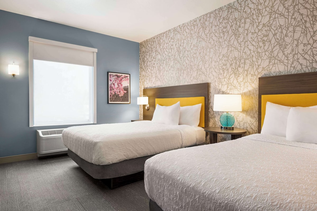 Hotelkamer met twee bedden, een raam met zonwering, een wandairconditioner en behang met een patroon.