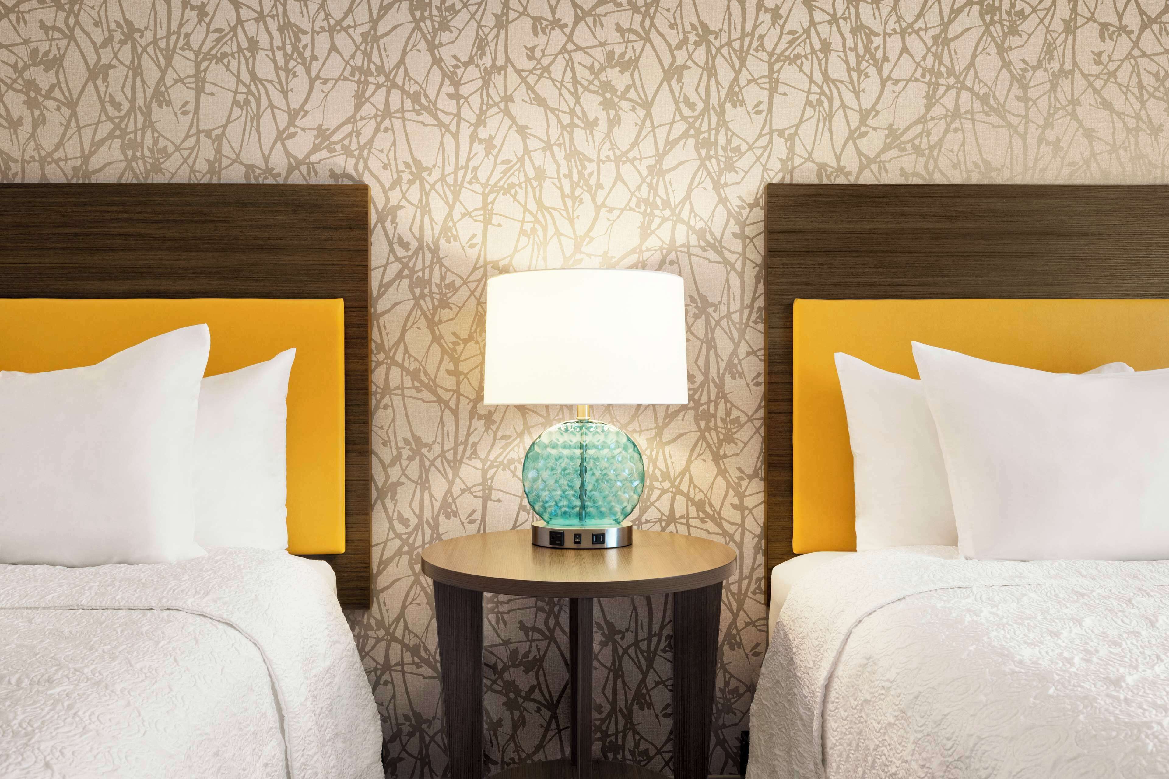 Une chambre d’hôtel avec deux lits, chacun doté d’une tête de lit jaune et de draps blancs. Une table ronde surmontée d’une lampe décorative bleue est placée au centre, entre les deux lits.