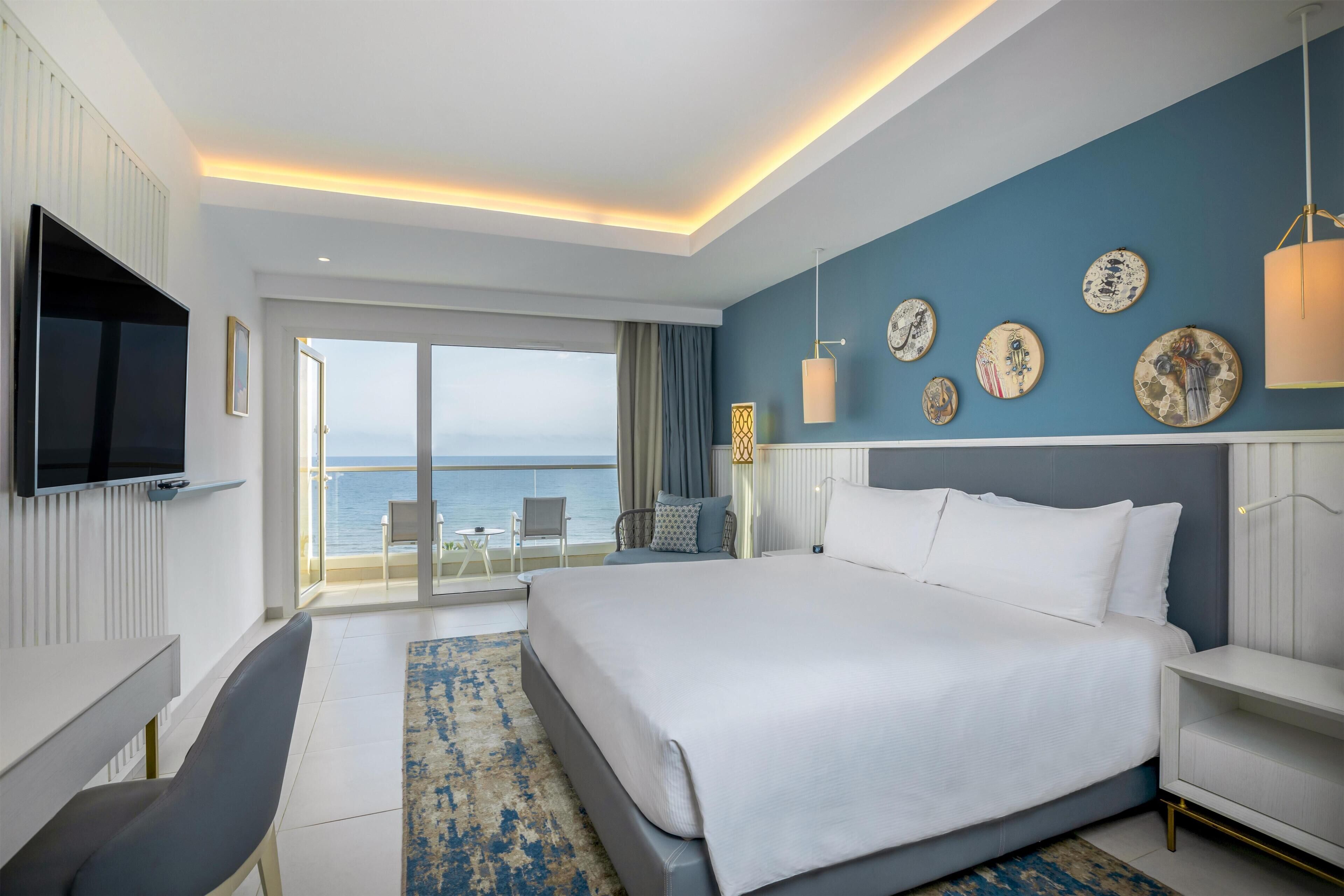 Una habitación de hotel moderna con una cama grande, un televisor, un balcón con vistas al mar y platos decorativos en la pared.