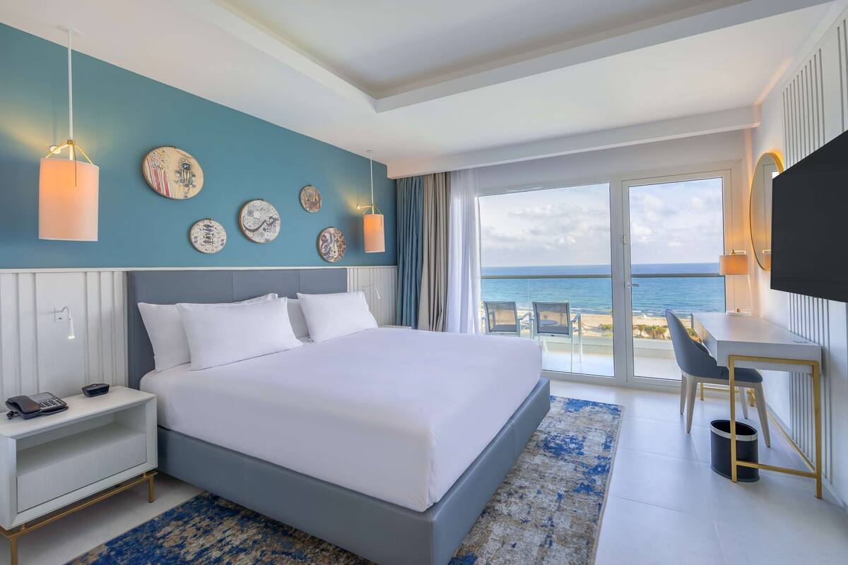 Una habitación de hotel con cama, mesita de noche, escritorio y un ventanal con vistas al mar.