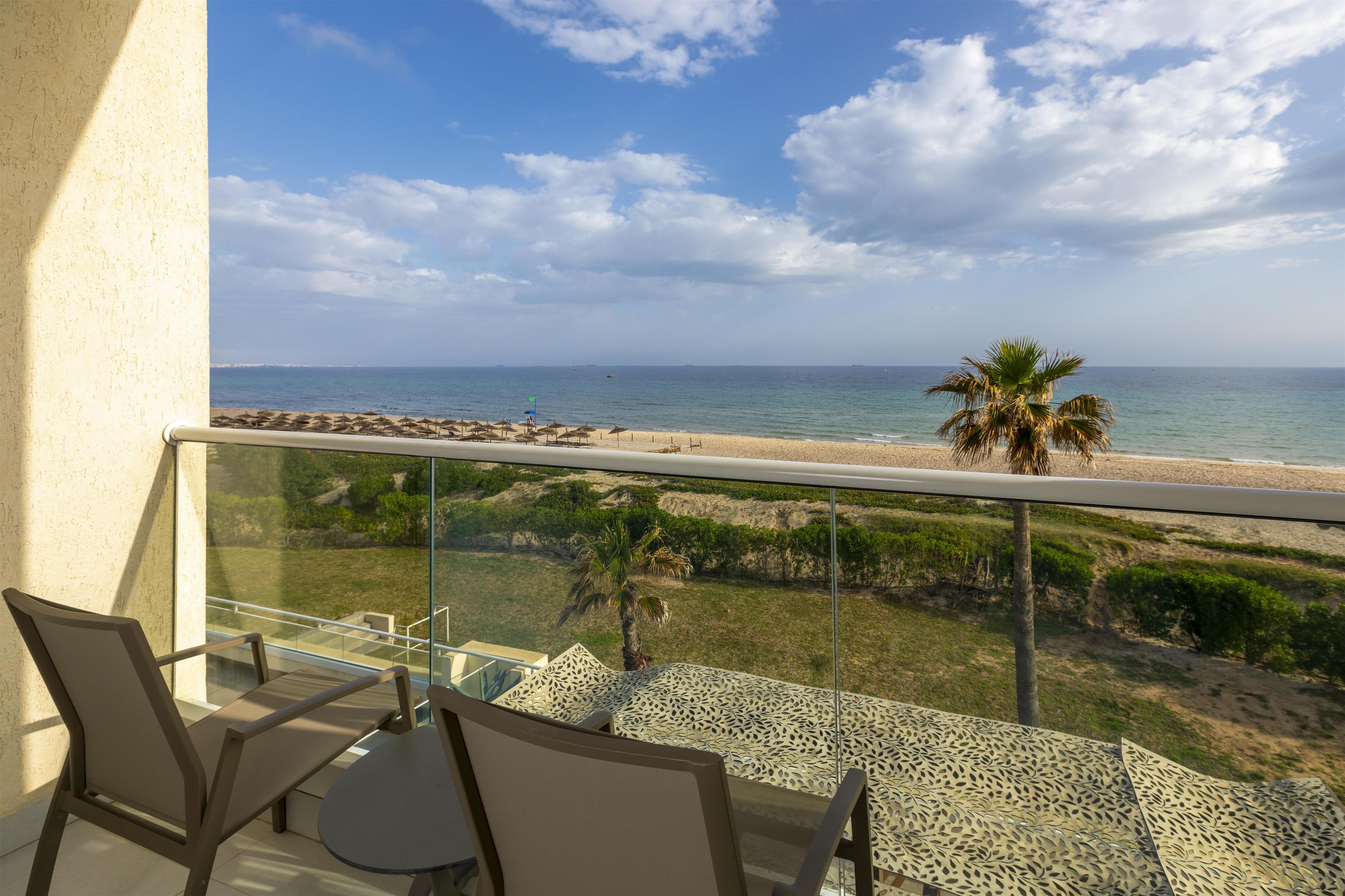 A balcony with a glass railing overlooking a beach, a palm tree, and a clear sky.