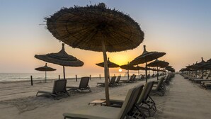 Une plage bordée de rangées de chaises longues et d’ombrelles en chaume, un ciel dégagé et l’océan en toile de fond.