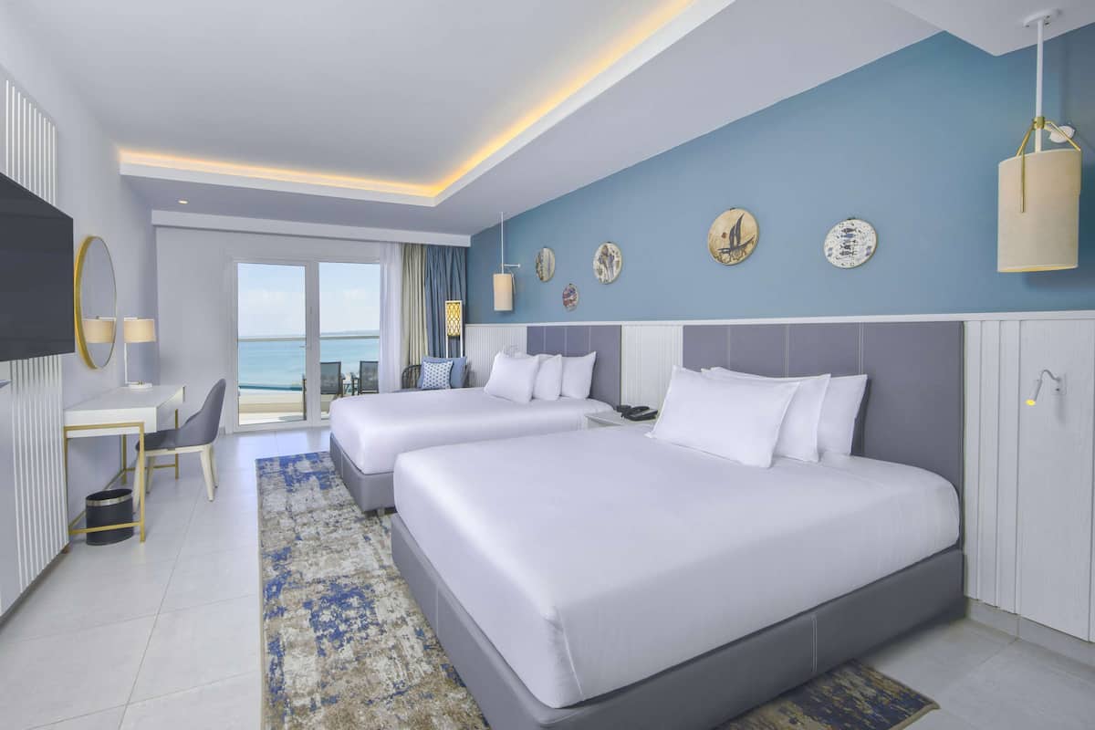 Habitación de hotel con dos camas, un escritorio, una silla, televisor y vista al mar.