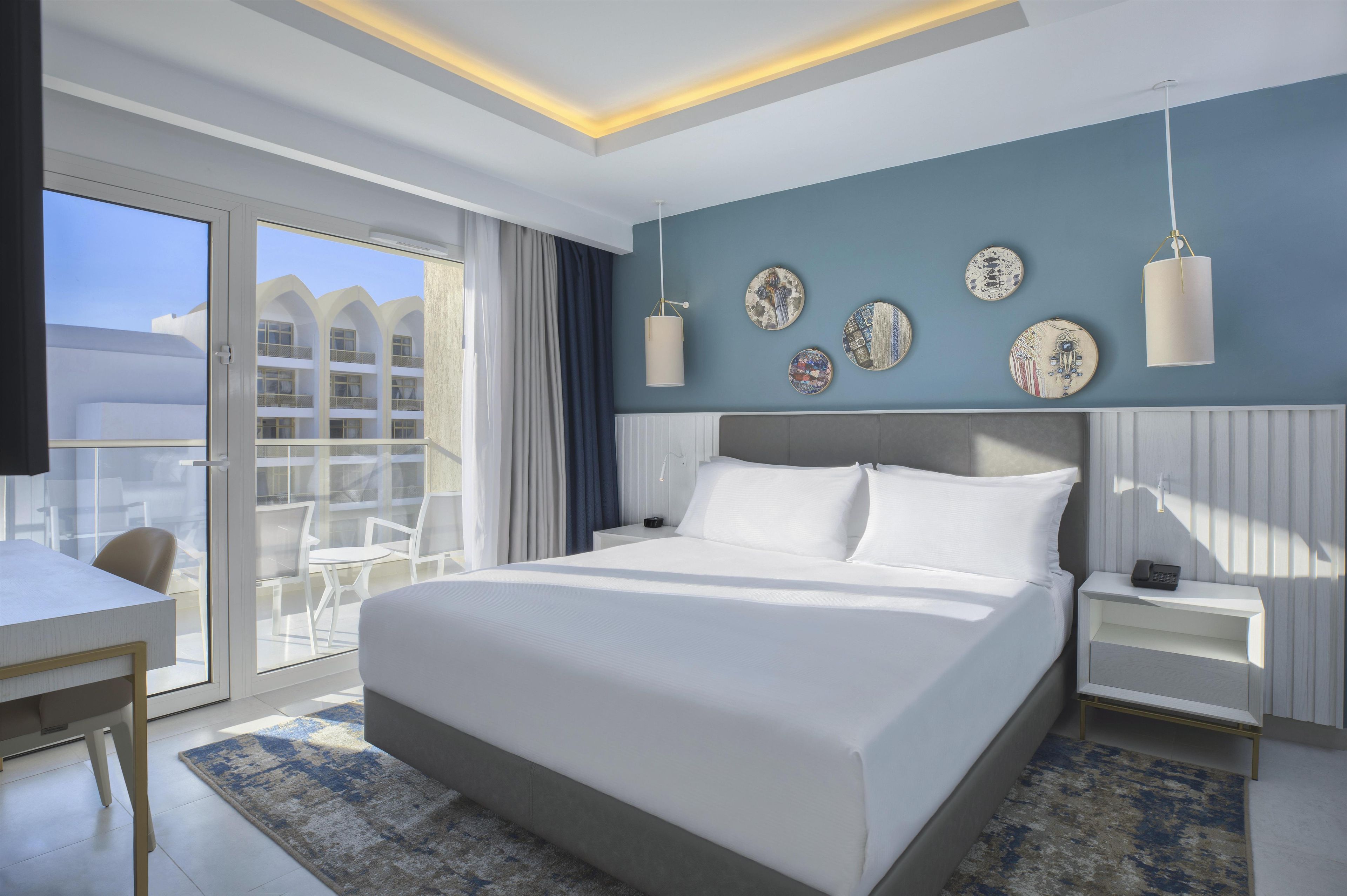 Una habitación de hotel moderna con una cama grande, un escritorio y un balcón con vista a un edificio.