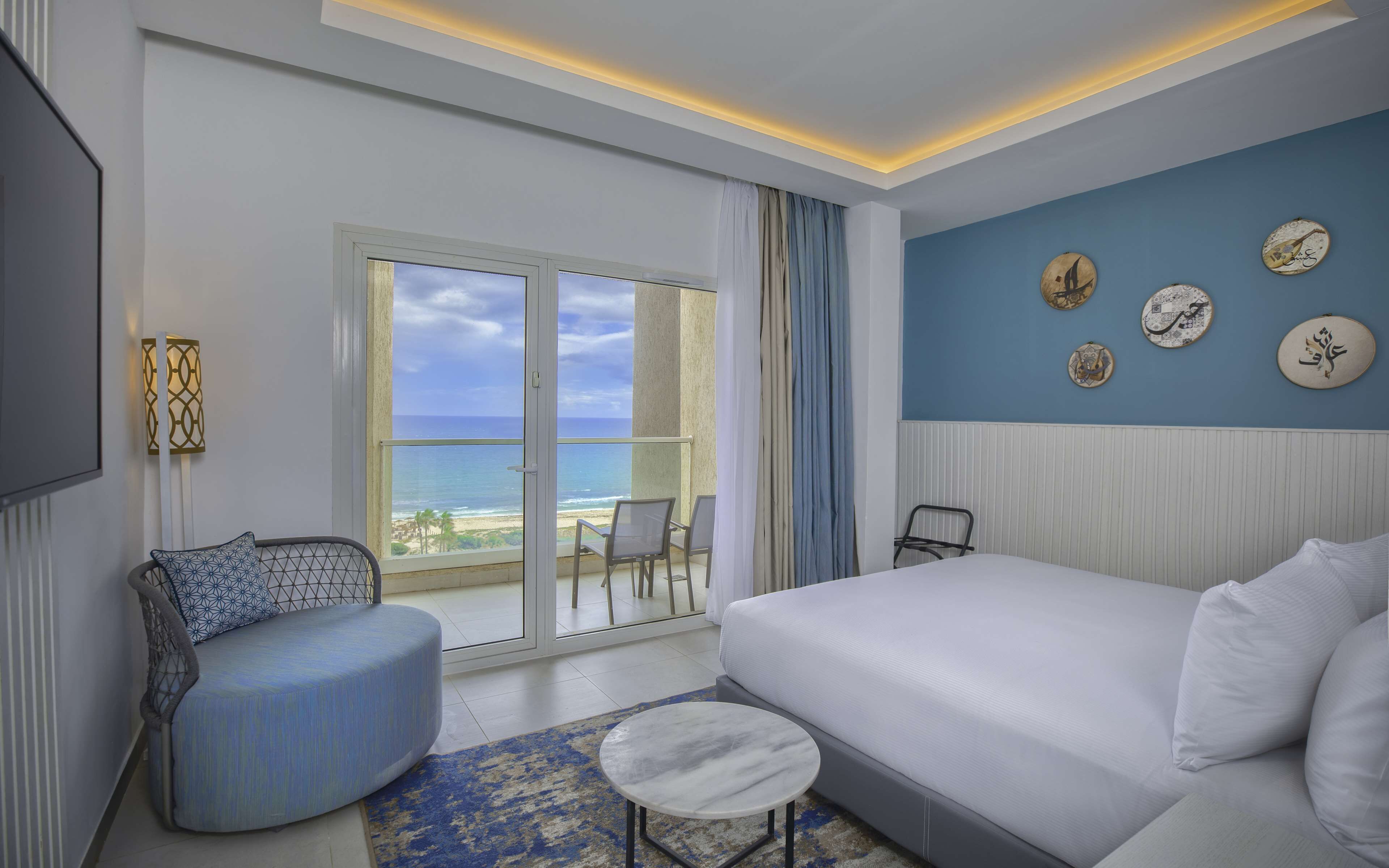 A hotel room with a large bed, a seating area with a chair and ottoman, a small round table, and a balcony with a view of the beach and ocean.