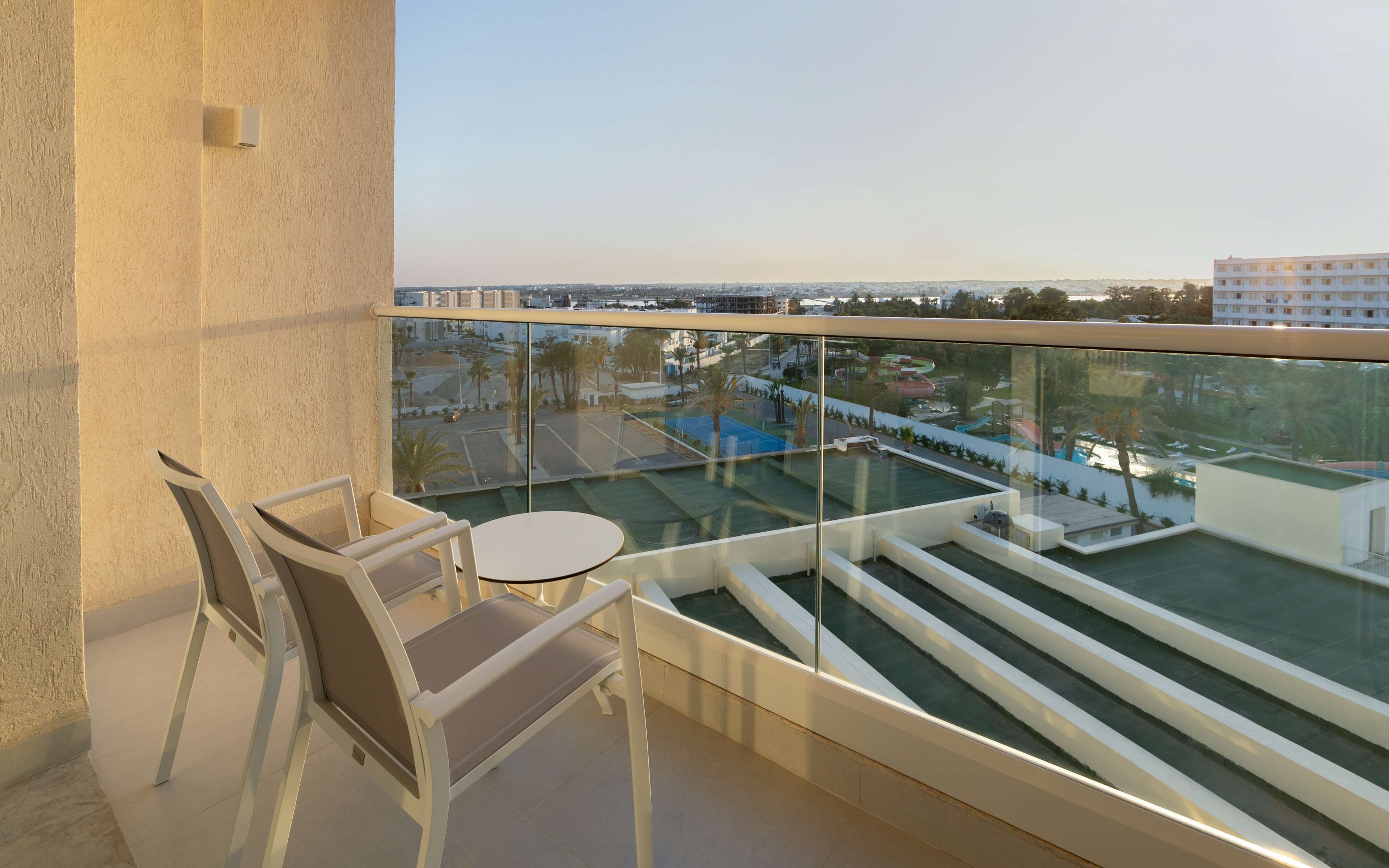 A balcony with a glass railing, two chairs, a small round table, and a view of a swimming pool and buildings.