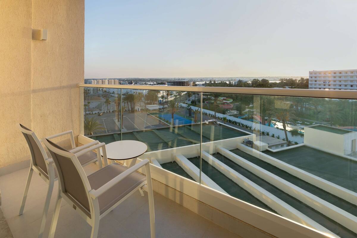 Un balcón con barandilla de vidrio, dos sillas, una mesita redonda y vistas a una piscina y edificios.