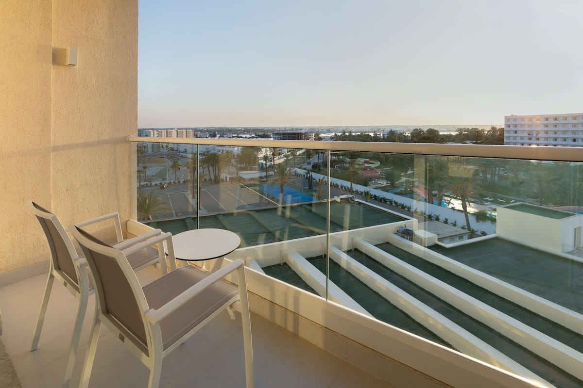 A balcony with a glass railing, two chairs, a small round table, and a view of a swimming pool and buildings.