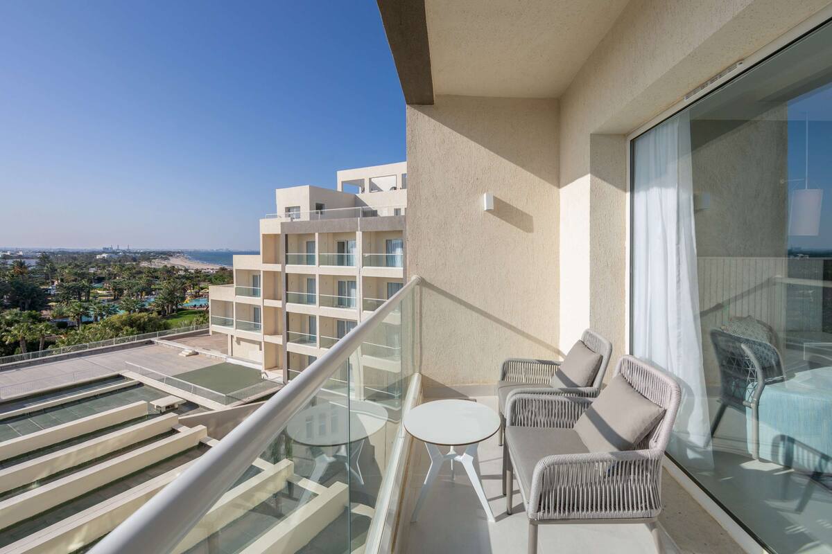 Un balcón con mobiliario de mimbre, una mesa de vidrio y vistas a la playa y edificios.