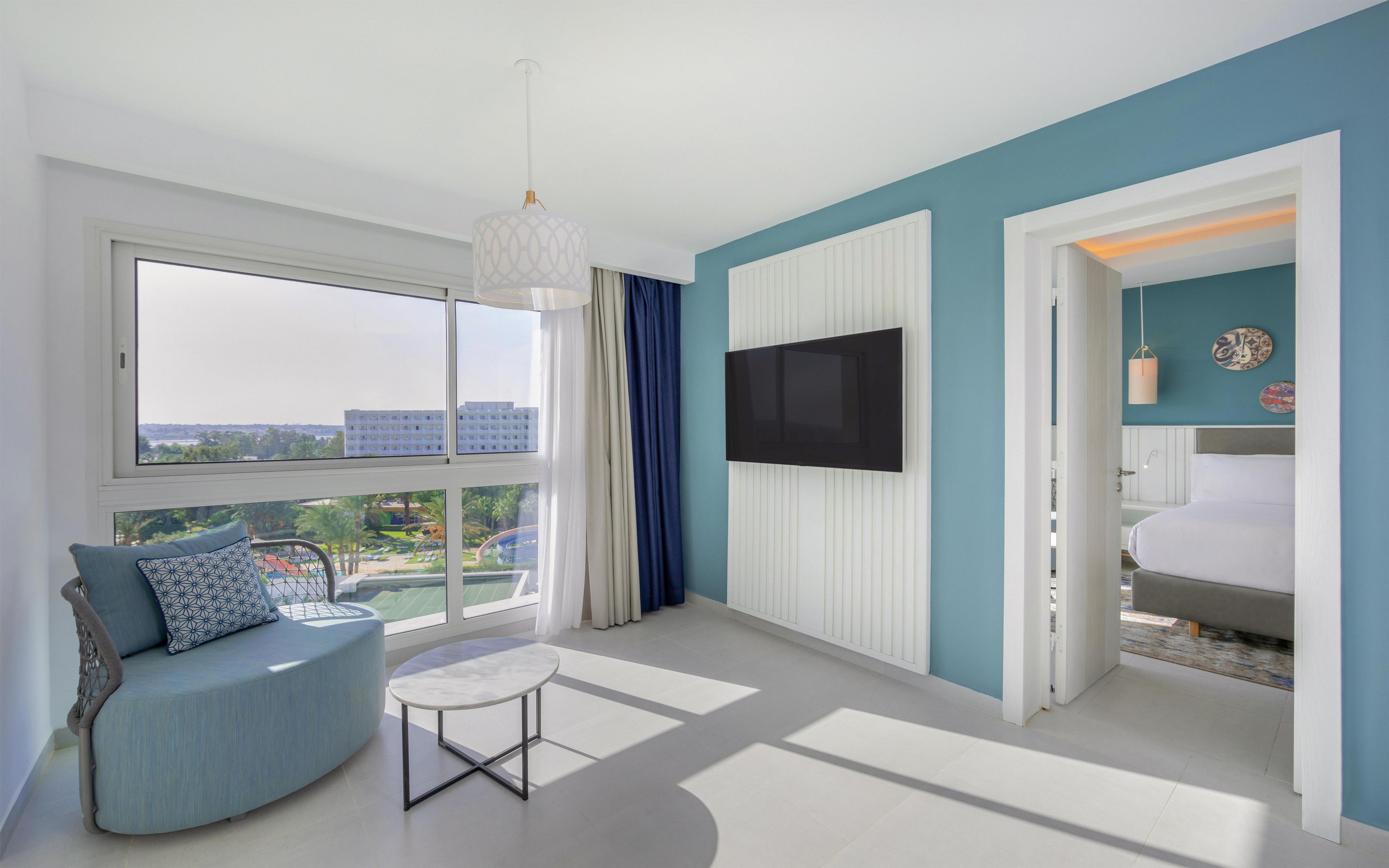 A modern hotel room with a large window, a sofa, a small table, and a television.