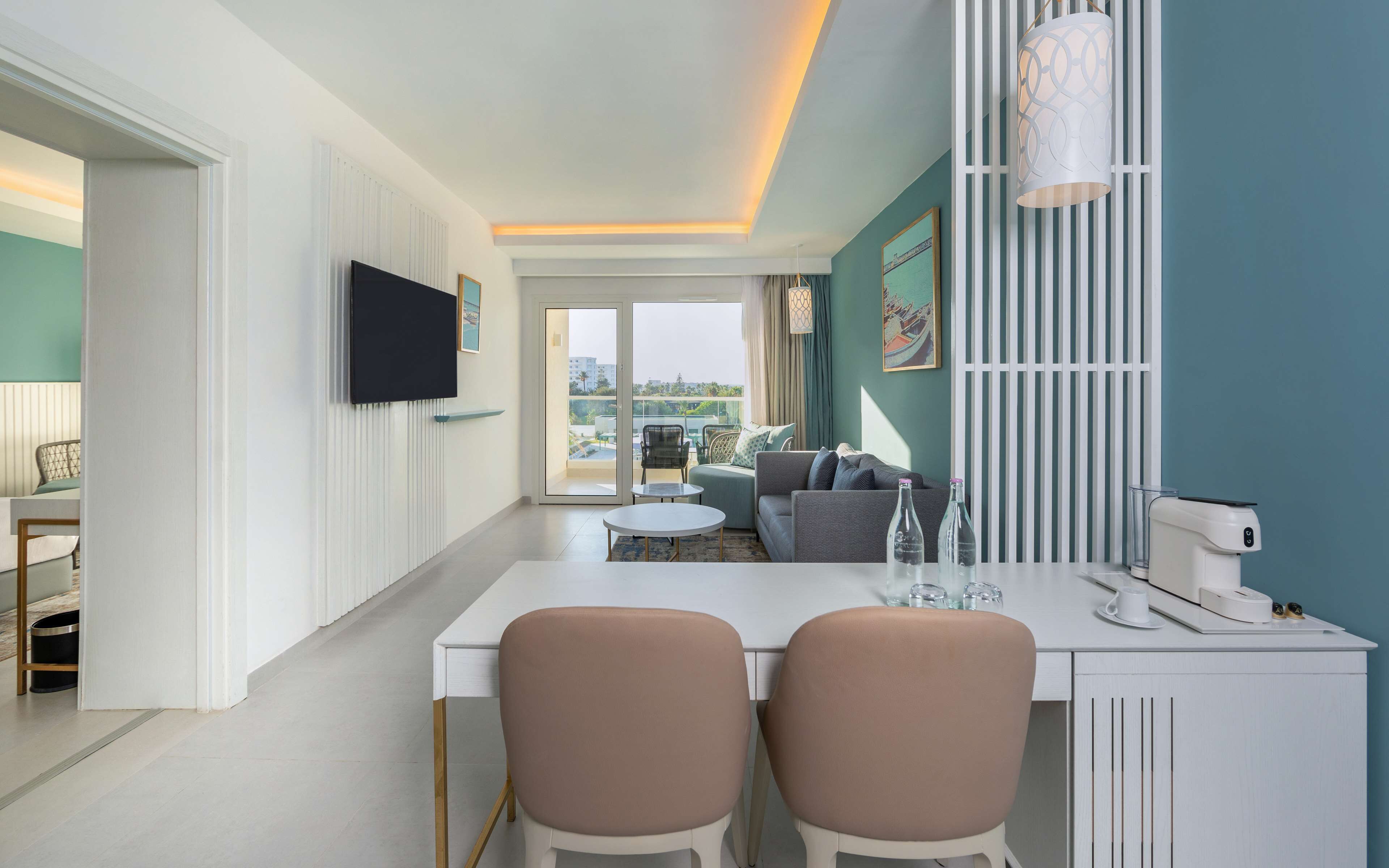Una sala de estar moderna con un sofá, una mesa de centro y un televisor montado en la pared. Se ve un balcón a través de la puerta de cristal.