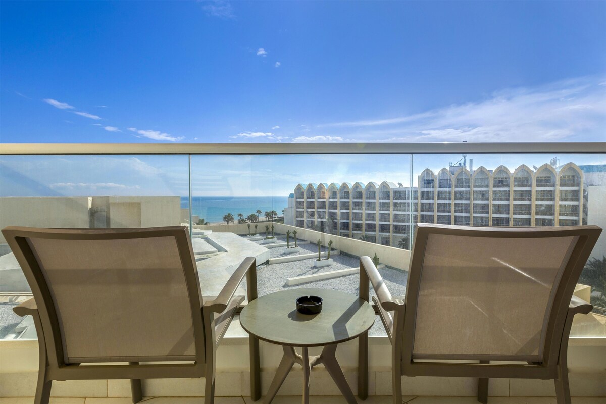 Un balcón con dos sillas y una mesa, con vistas a una playa y un edificio de hotel.