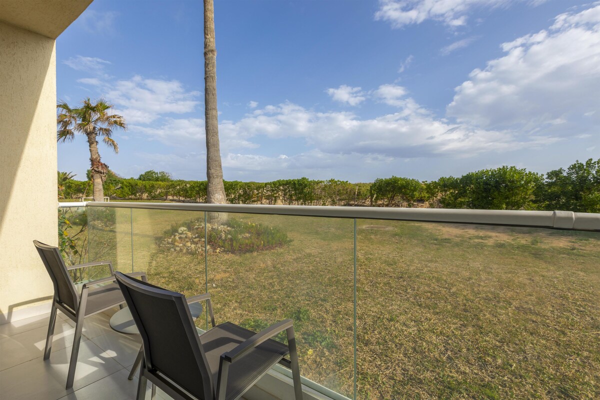 Un balcón con barandilla de vidrio, una palmera y vista a una zona verde.
