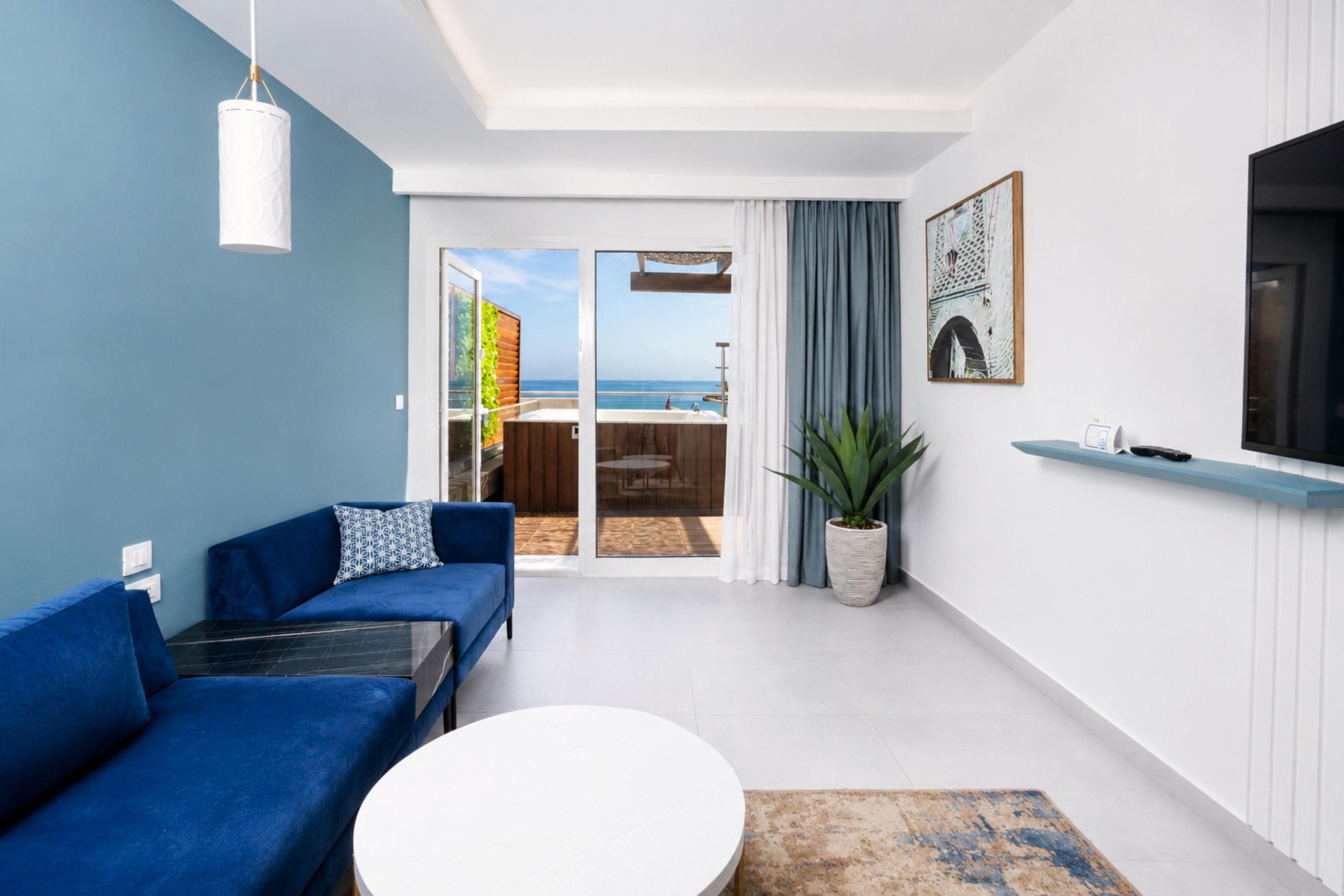 A modern living room with a blue sofa, a white coffee table, and a view of the sea through a glass door.