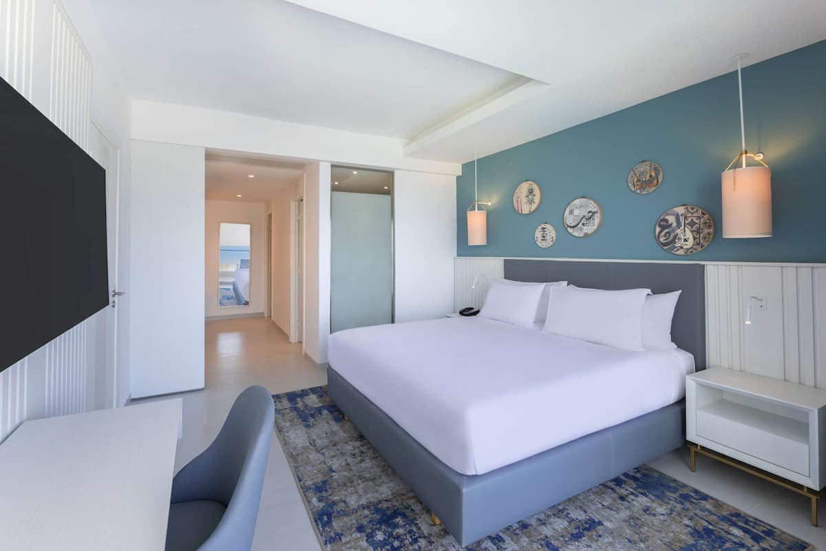 Un dormitorio moderno con una cama grande, un televisor de pantalla plana, un escritorio y vista al mar a través de una puerta.
