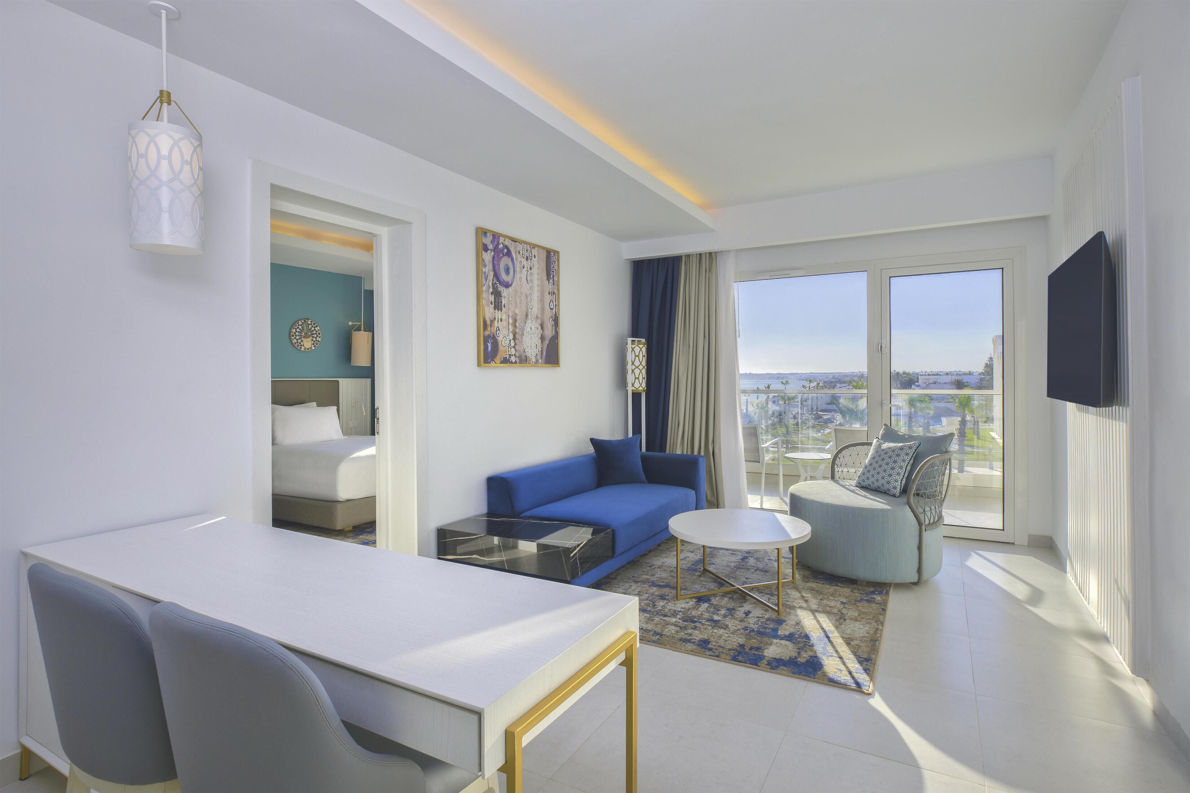 A modern hotel room with a dining area, a sofa, a chair, and a large window with a city view.