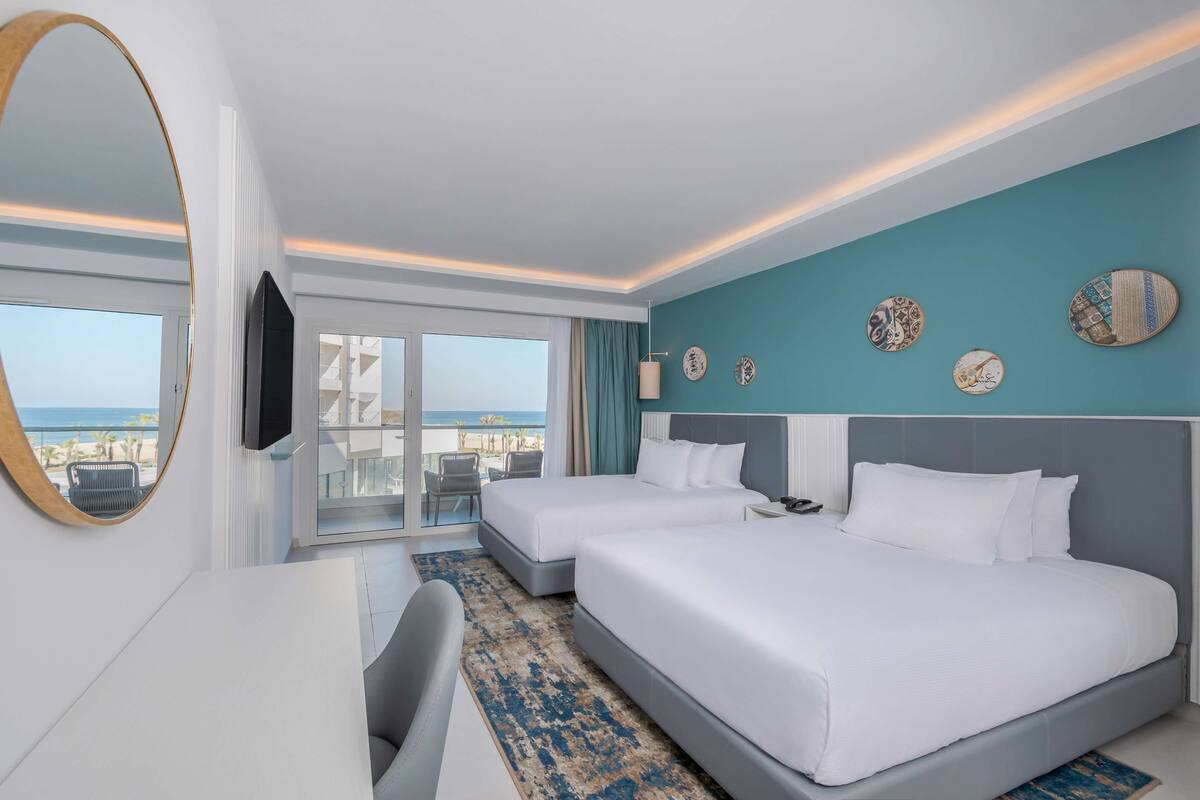 Habitación de hotel con dos camas, un espejo grande, televisión y balcón con vista a la playa.