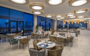 Een modern restaurant met ronde tafels en rieten stoelen, grote ramen met uitzicht op de oceaan en een plafond met ronde lampen.