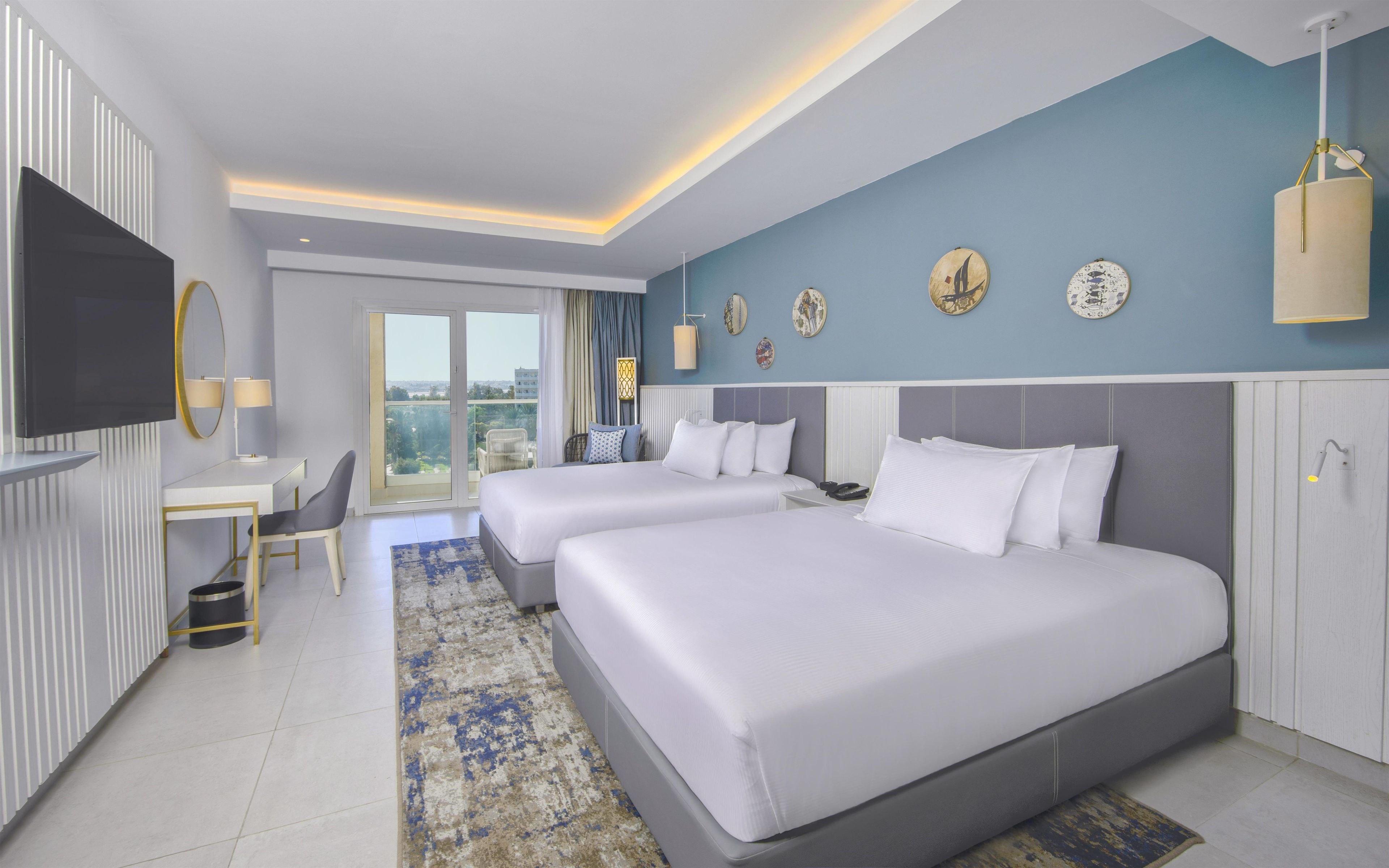 A hotel room with two beds, a desk, a TV, and a balcony with a view.