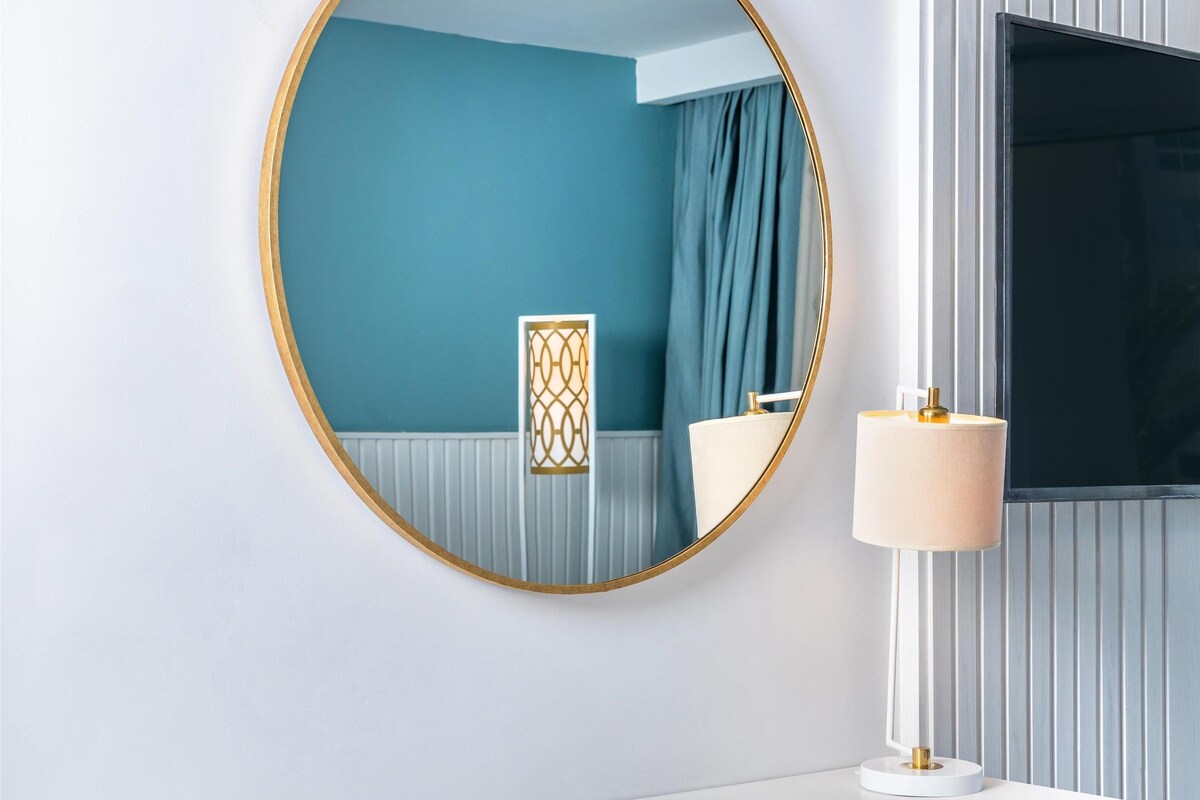Una habitación de hotel moderna con un escritorio blanco, una silla azul, un espejo redondo con marco dorado y una lámpara de noche.