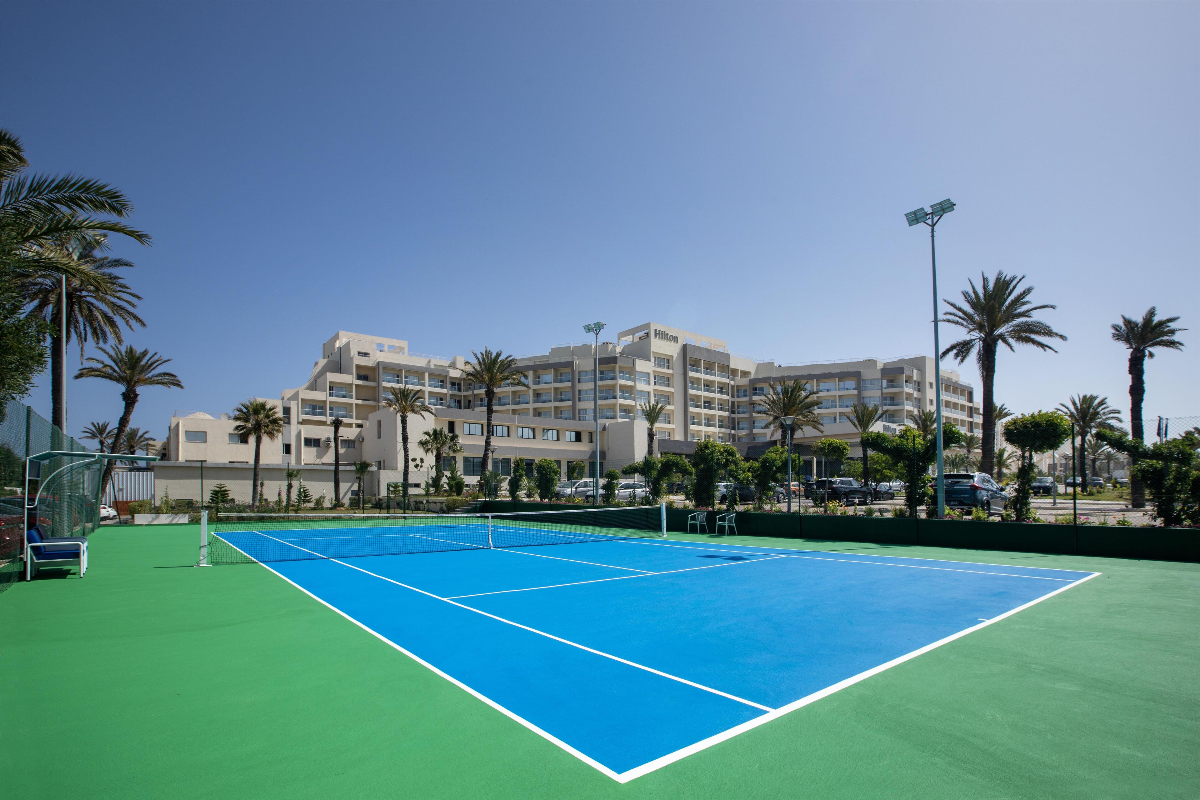 Una cancha de tenis con una red, rodeada de palmeras y un edificio de hotel al fondo.