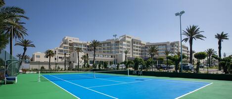 Una cancha de tenis con una red, rodeada de palmeras y un edificio de hotel al fondo.