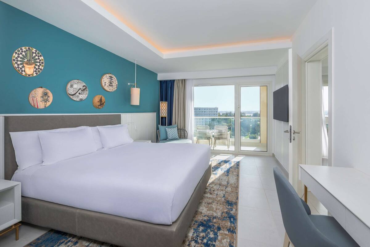 Ett modernt hotellrum med en stor säng, en platt-TV, en balkong med utsikt över staden och dekorativ väggkonst.