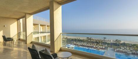 A balcony with a view of a pool and beach, featuring chairs and a table.