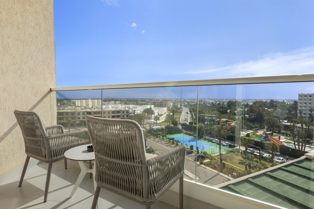 Un balcón con sillas de mimbre y una mesa, con vistas a una piscina y edificios.