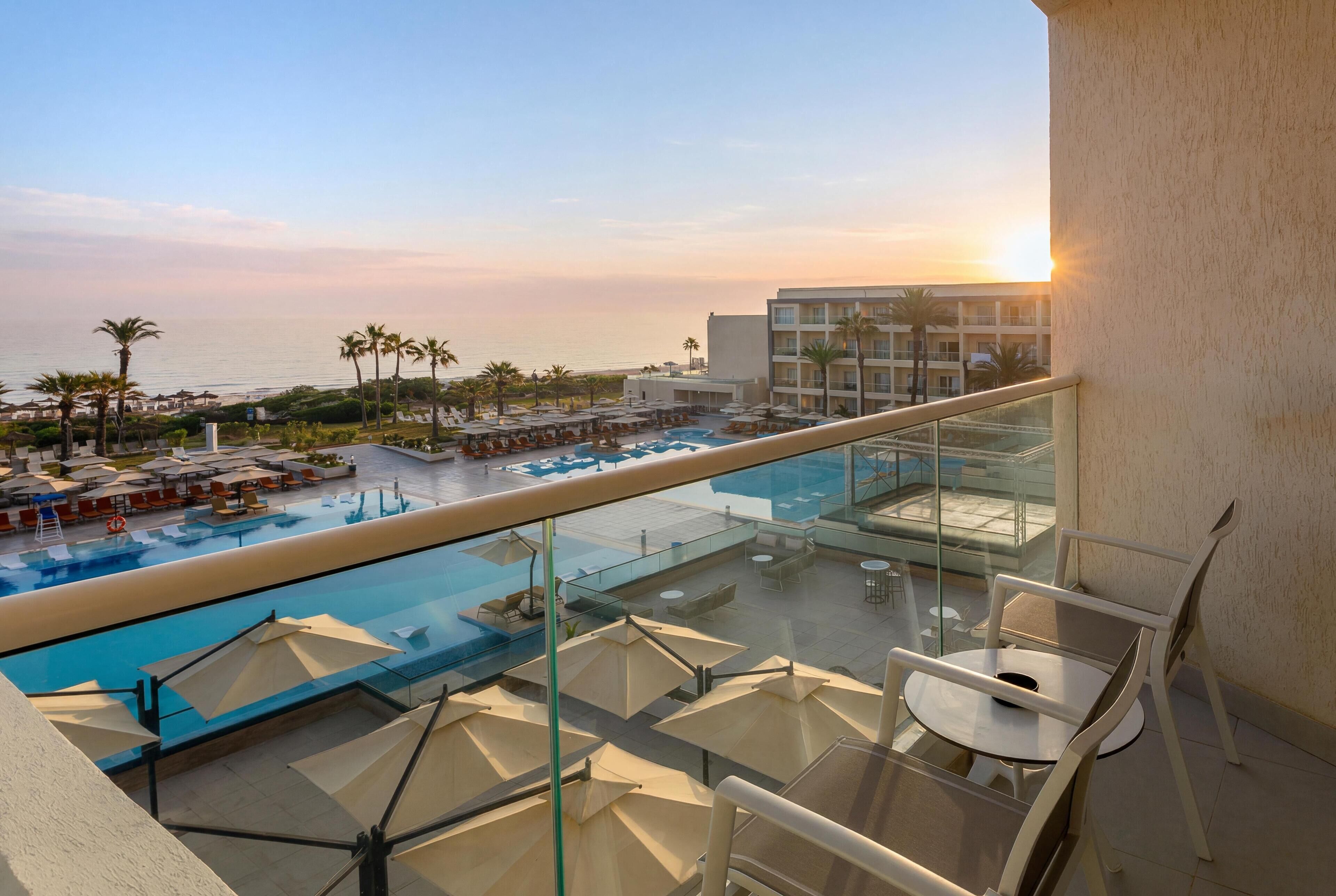 A rooftop pool area with lounge chairs and umbrellas, overlooking a pool and a building with balconies.
