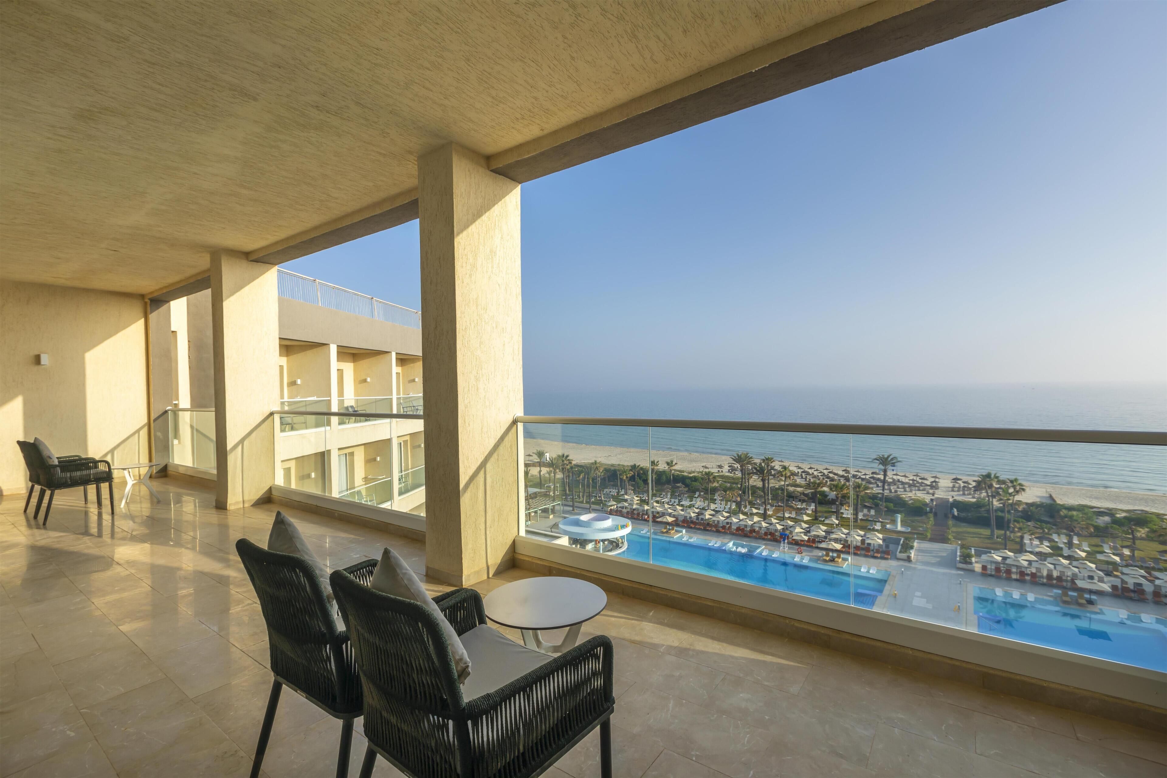 Un balcon avec vue sur une piscine et une plage, comprenant des chaises et une table.