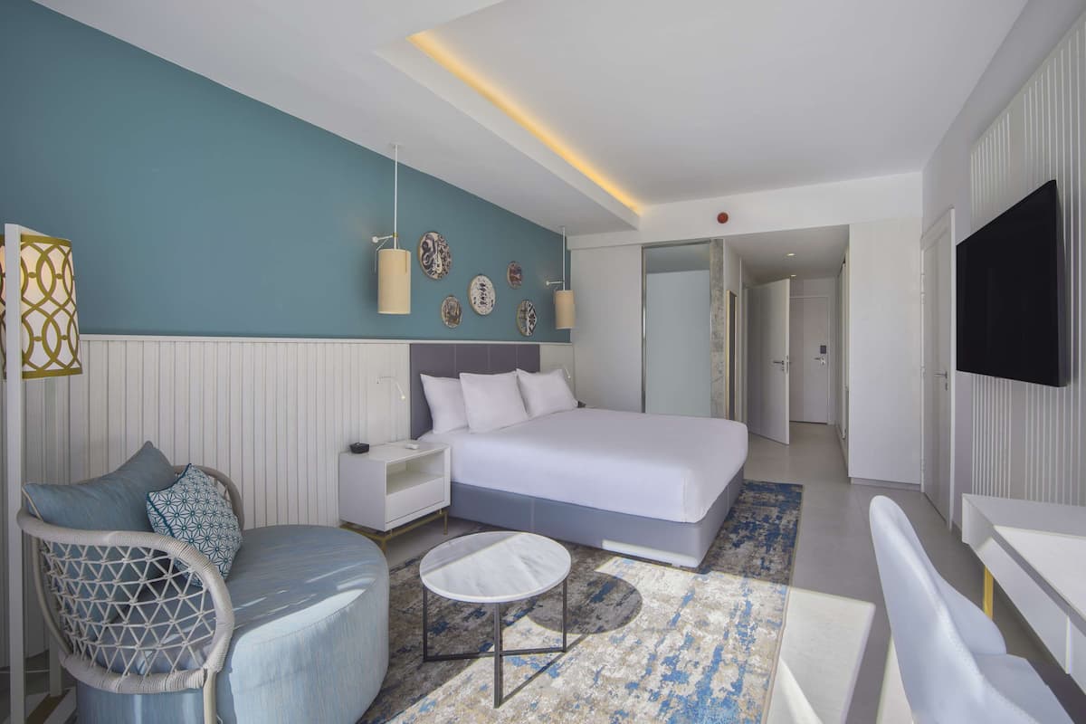 Una habitación de hotel moderna con una cama grande, una zona de estar con una silla y una mesita pequeña, un televisor montado en la pared y una decoración mural con varios objetos enmarcados.
