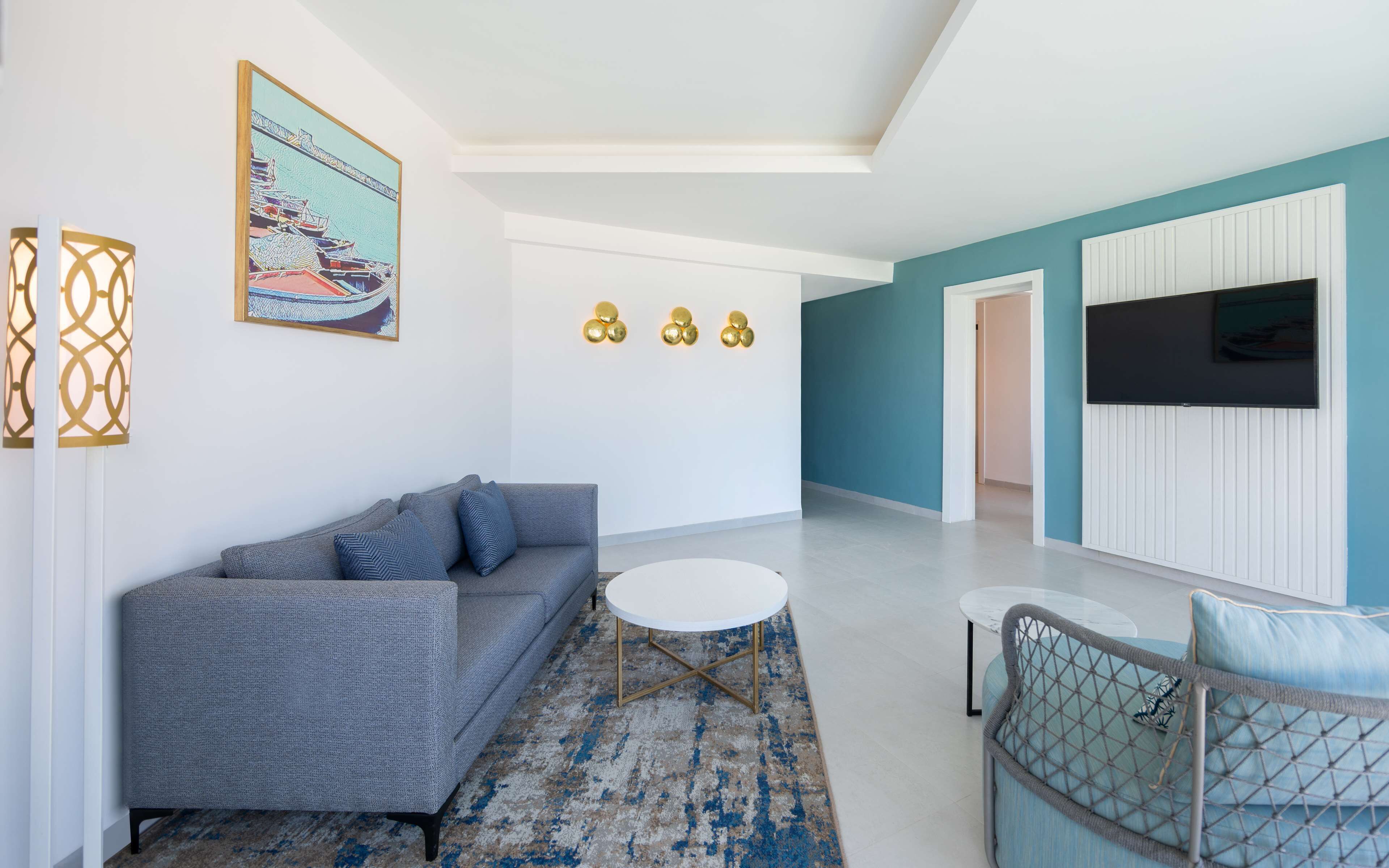 A modern living room with a grey sofa, a round coffee table, and a blue patterned rug. There is a painting of boats on the wall and a flat-screen TV mounted on the wall.