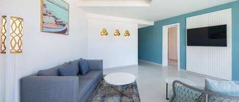 A modern living room with a grey sofa, a round coffee table, and a blue patterned rug. There is a painting of boats on the wall and a flat-screen TV mounted on the wall.