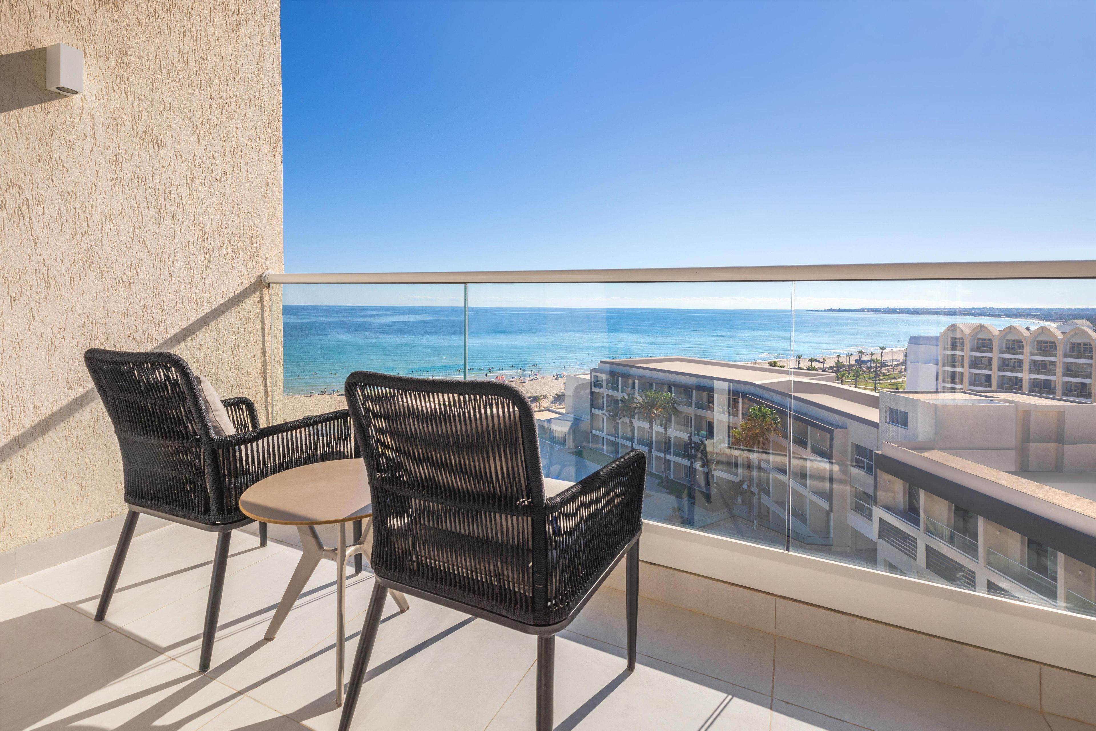 A balcony with two wicker chairs and a small table, overlooking a beach and a modern building.