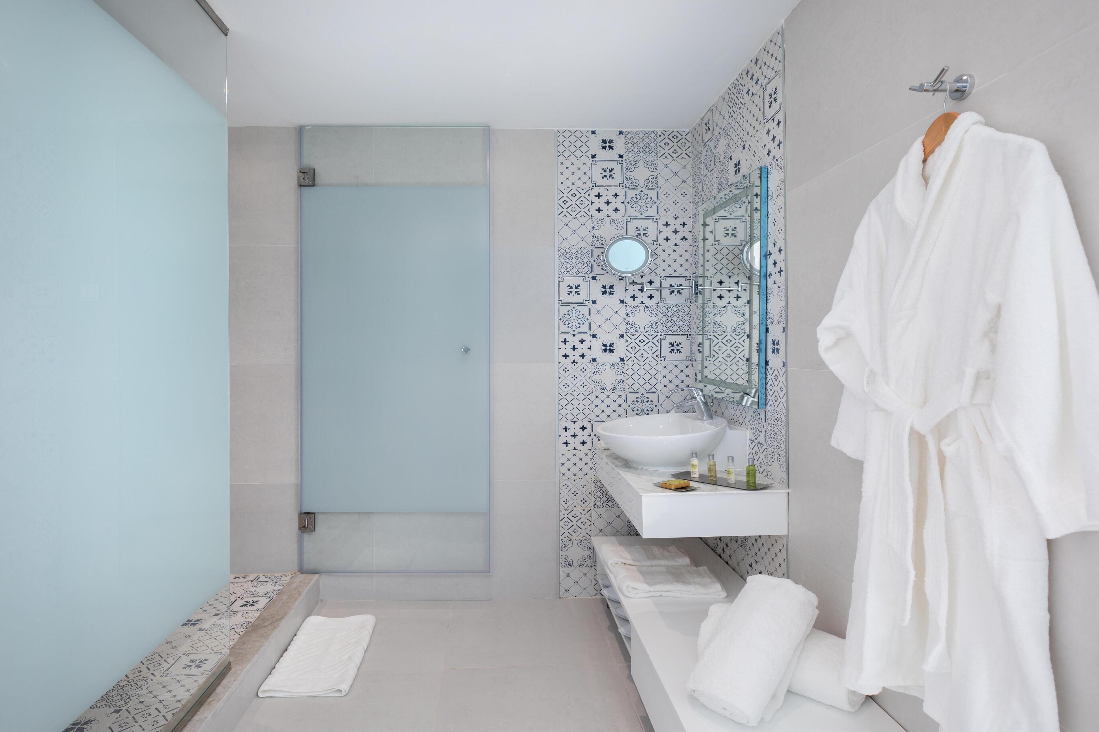A modern bathroom with a walk-in shower, a large mirror, a white sink, and a white bathrobe hanging on the wall.