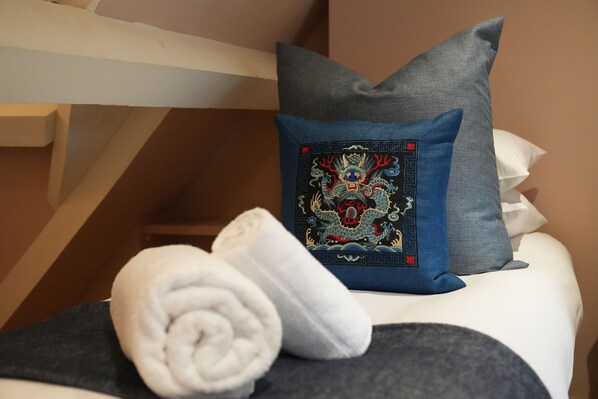 Un lit avec un oreiller gris et un oreiller bleu orné d’un motif de dragon, une serviette blanche roulée et une couverture grise.
